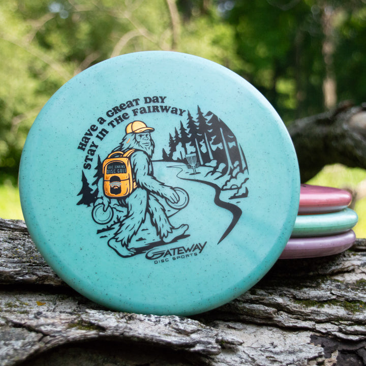 A turquoise Eric Jenkins Signature Series Chief - Diamond Hemp putter shows Bigfoot with a disc bag and the phrase "Have a Great Day Stay in the Fairway," relaxing on a log outdoors.