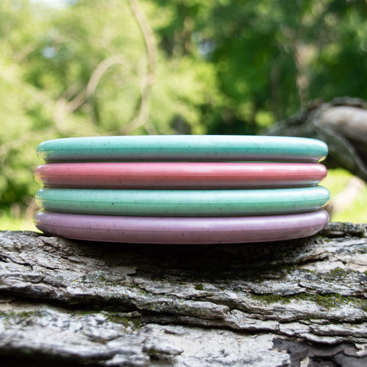 Four pastel-colored Eric Jenkins Signature Series Chief - Diamond Hemp discs—two pink, one blue, and one purple—resembling low profile putters, are stacked on a tree trunk outdoors with green foliage in the background.