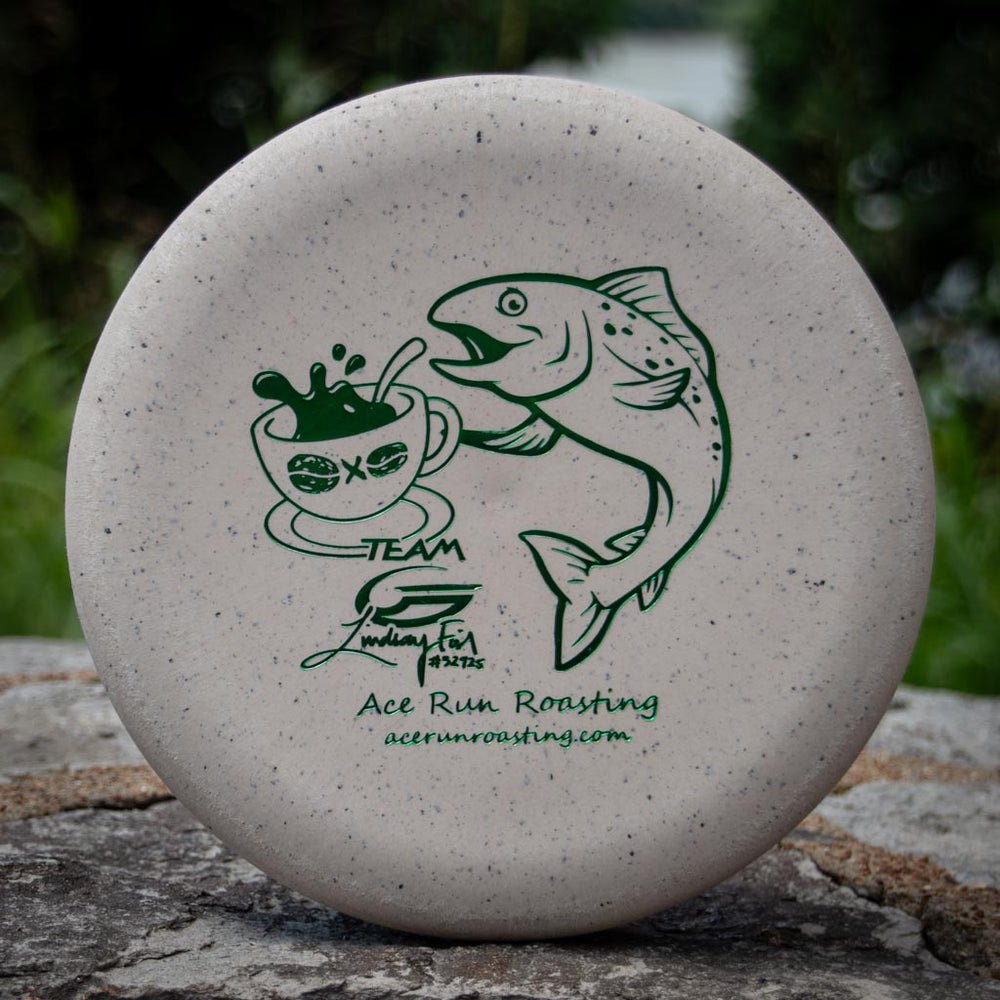 The Lindsay Fish Warlock - Coffee Blend features a white disc golf disc with a green fish and coffee cup graphic, "Ace Run Roasting," and "acerrunroasting.com"—sales support the Lindsay Fish initiative.