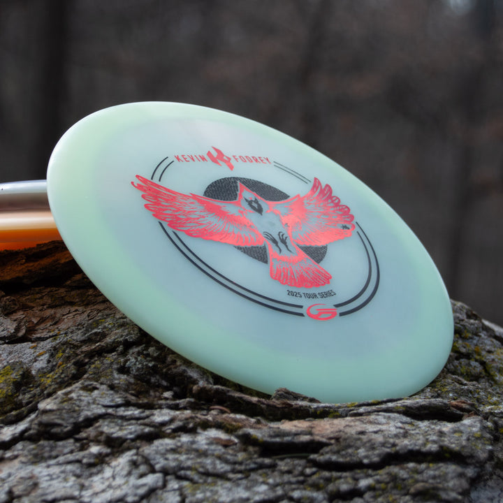 Kevin Fodrey's Spell - Superglow, an overstable driver disc with a red bird design and "Kevin Jones, 2023 Tour Series" text, is displayed outdoors on rough tree bark.