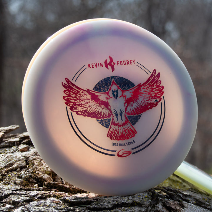 The Kevin Fodrey's Spell - Superglow is an overstable driver featuring a red bird graphic and "Kevin Fodrey's 2025 Tour Series" text, pictured from the NXT Color Glow line resting on a tree trunk outdoors.