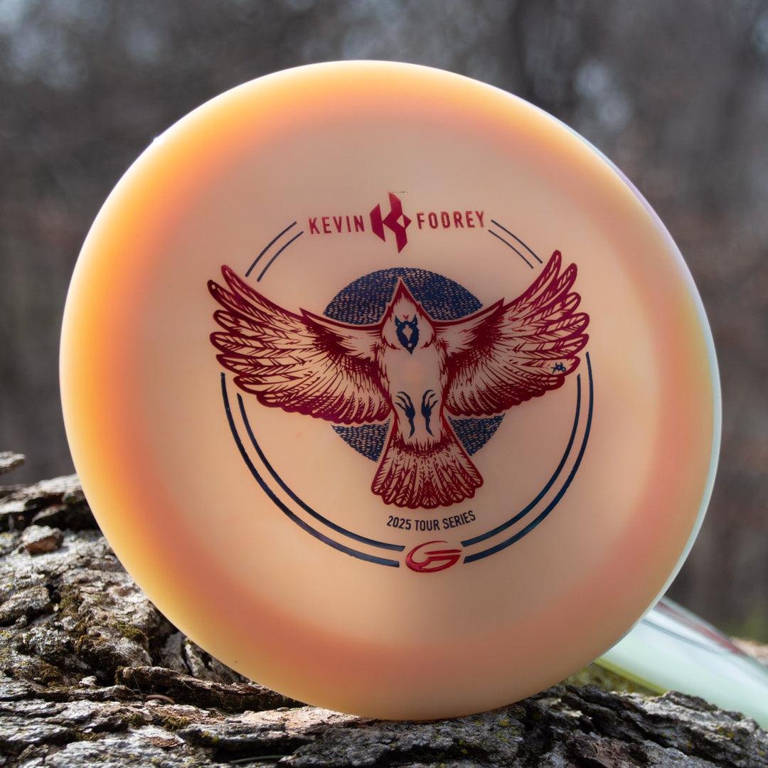 Close-up of Kevin Fodrey's Spell - Superglow driver with a red eagle graphic, "Kevin Fodrey," and "2025 Tour Series" text, resting on a tree trunk outdoors.