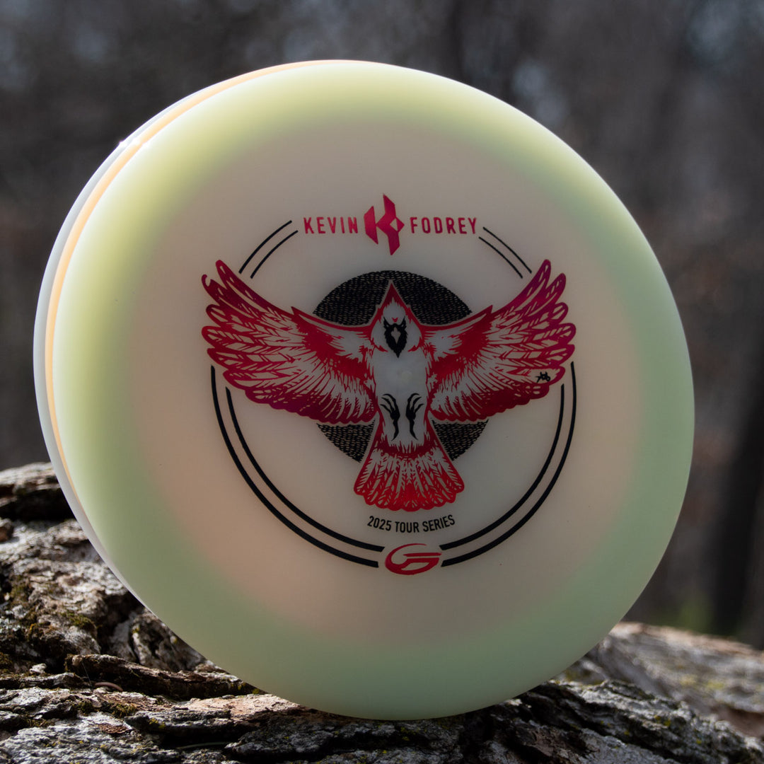 The Kevin Fodrey's Spell - Superglow disc, with a red and black bird graphic and "Kevin Fodrey 2023 Tour Series" marking, rests against a tree trunk—crafted in NXT Color Glow plastic for stylish outdoor play.
