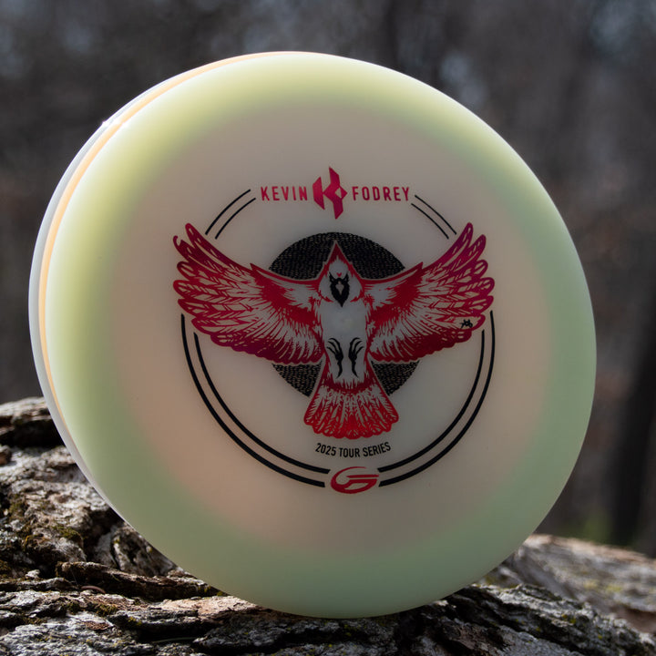 The Kevin Fodrey's Spell - Superglow disc, with a red and black bird graphic and "Kevin Fodrey 2023 Tour Series" marking, rests against a tree trunk—crafted in NXT Color Glow plastic for stylish outdoor play.
