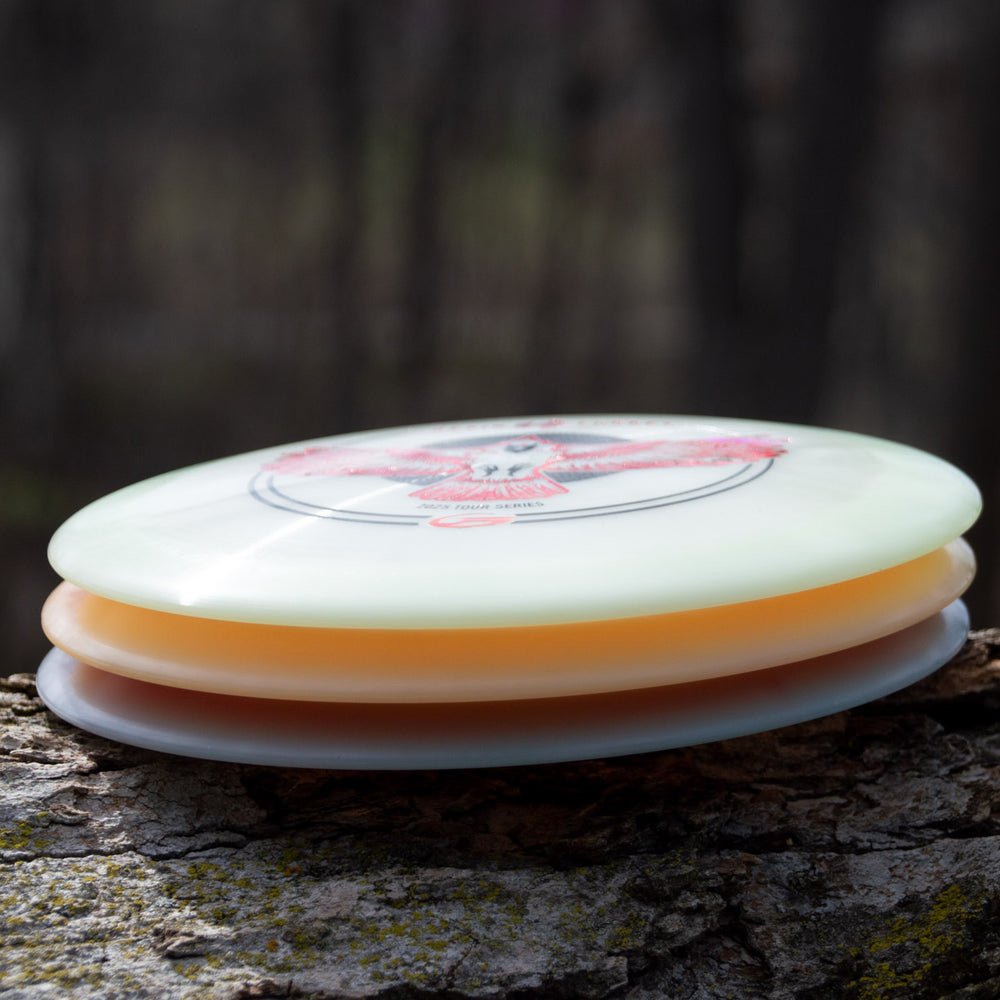 Three disc golf discs, including Kevin Fodrey's Spell - Superglow overstable driver, stacked on a tree trunk with a blurred forest background.