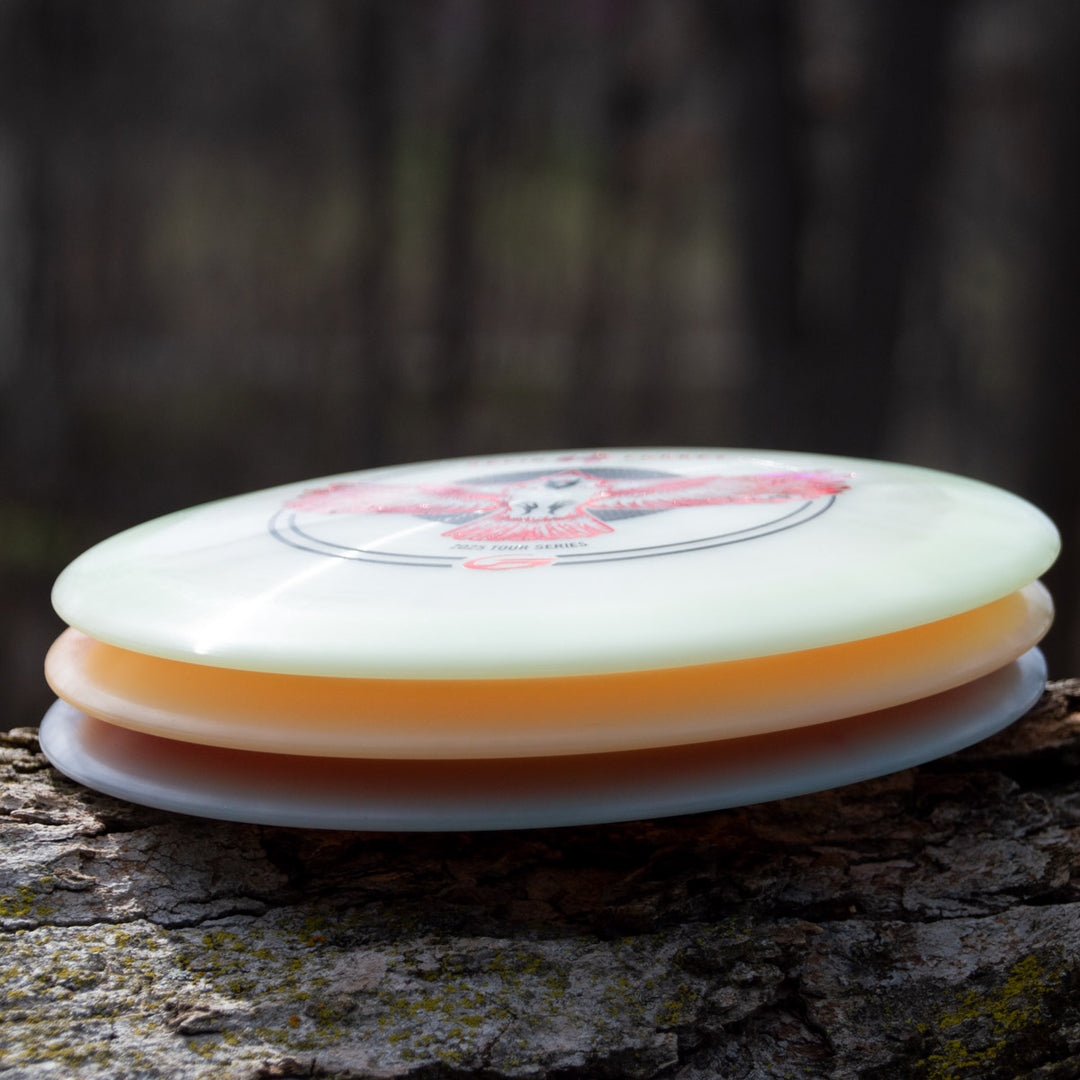 Three disc golf discs, including Kevin Fodrey's Spell - Superglow overstable driver, stacked on a tree trunk with a blurred forest background.