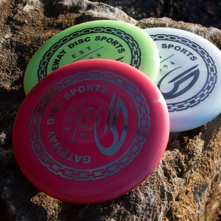 The Gateway Disc Sports Wizard - Evolution - Frosty discs in red, green, and white sit on a large rock, their logos and text brightly illuminated by sunlight.