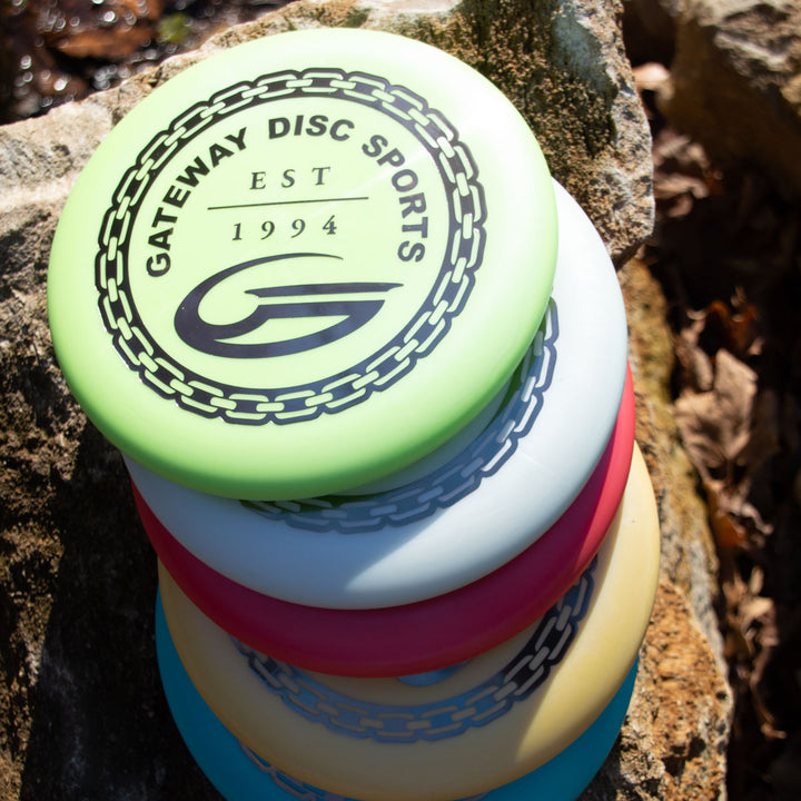 A stack of colorful disc golf discs rests on a rock outdoors, with the top one showing the "Gateway Disc Sports" logo and "Est 1994," featuring the iconic Wizard - Evolution - Frosty model.