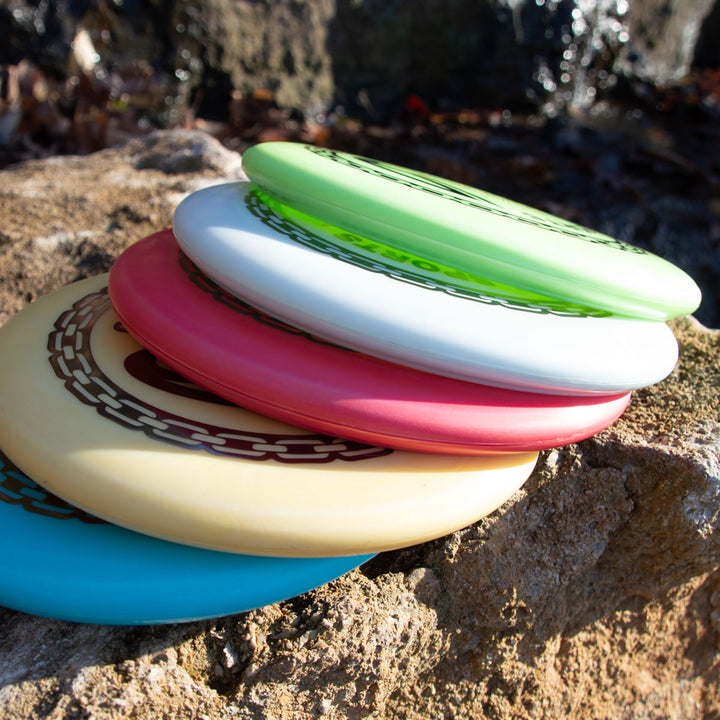 Five Wizard - Evolution - Frosty disc golf discs are stacked on a rock outdoors, sunlight highlighting their vibrant colors and unique chain designs.