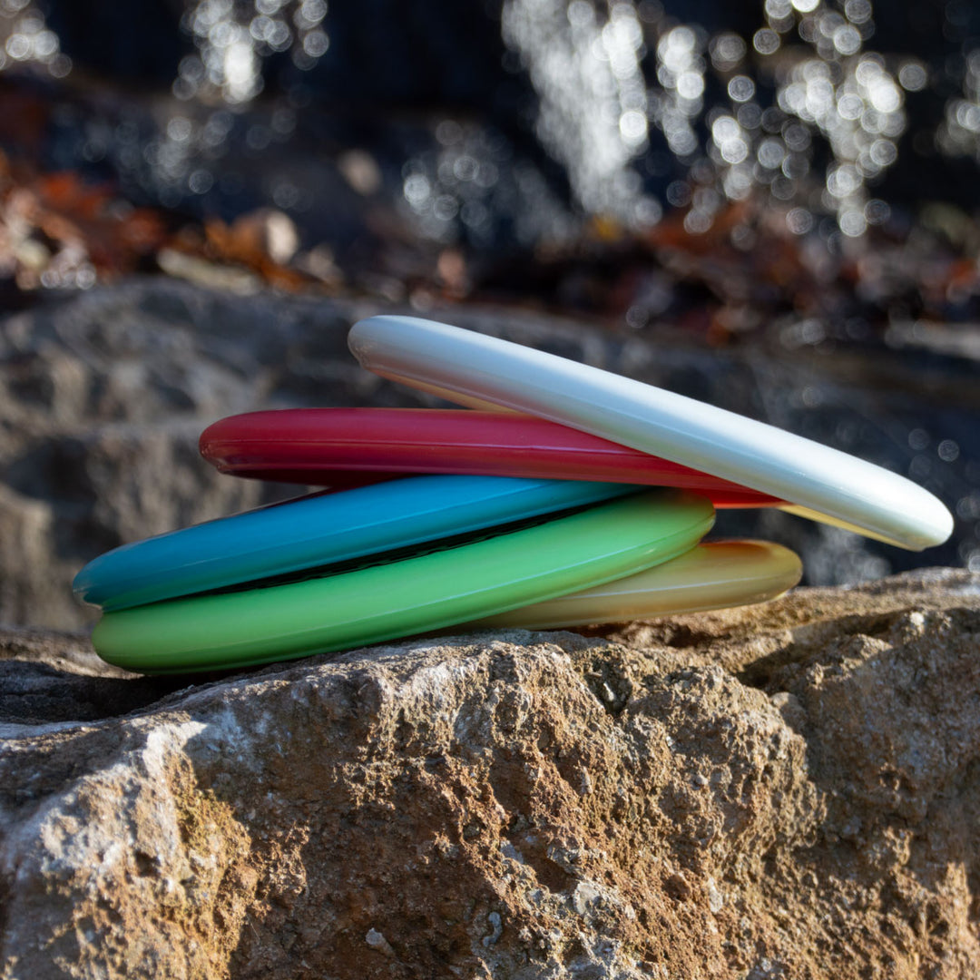 A stack of colorful Wizard - Evolution - Frosty discs in white, red, blue, green, and yellow rests on a large rock outdoors with a blurred water background.