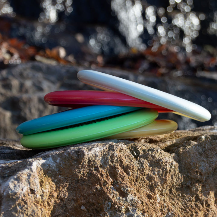 A stack of colorful Wizard - Evolution - Frosty discs in white, red, blue, green, and yellow rests on a large rock outdoors with a blurred water background.