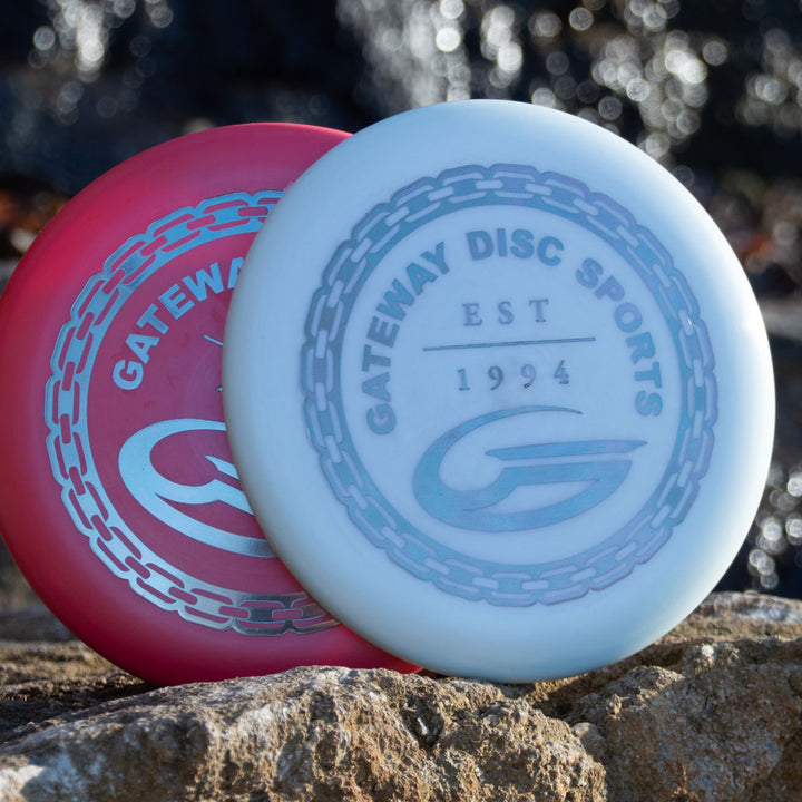 A red Gateway disc and a white Frosty Wizard - Evolution disc from Gateway Disc Sports rest on a rock against an outdoor backdrop.
