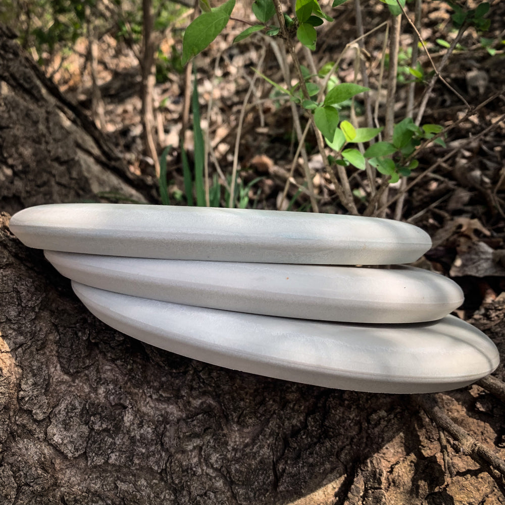 Three Gavin Rathbun's Retro Warlock - Nylon Firm discs in gray are stacked on a tree trunk outdoors, surrounded by green leaves and branches in the background.