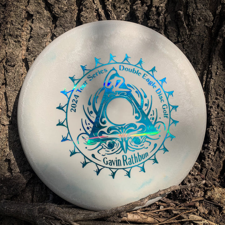 A white Gavin Rathbun's Retro Warlock - Nylon Firm approach disc from the Retro Series, with blue holographic "2024 Tour Series Double Eagle Disc Golf Gavin Rathbun" text, rests against tree bark.