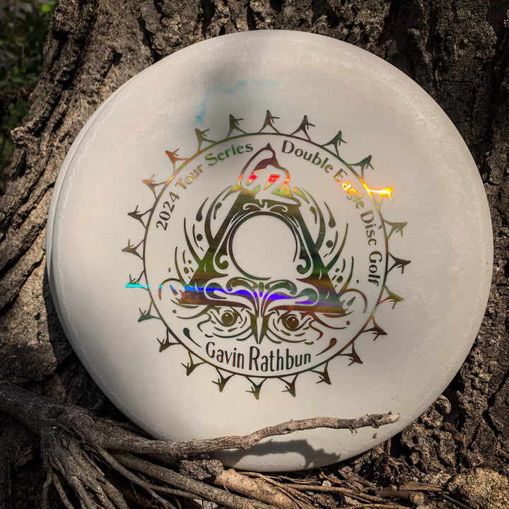 Gavin Rathbun's Retro Warlock - Nylon Firm, a white disc with "2024 Tour Series Double Eagle Disc Golf" and his name in decorative lettering, rests against tree bark—ideal for fans of quality approach discs.