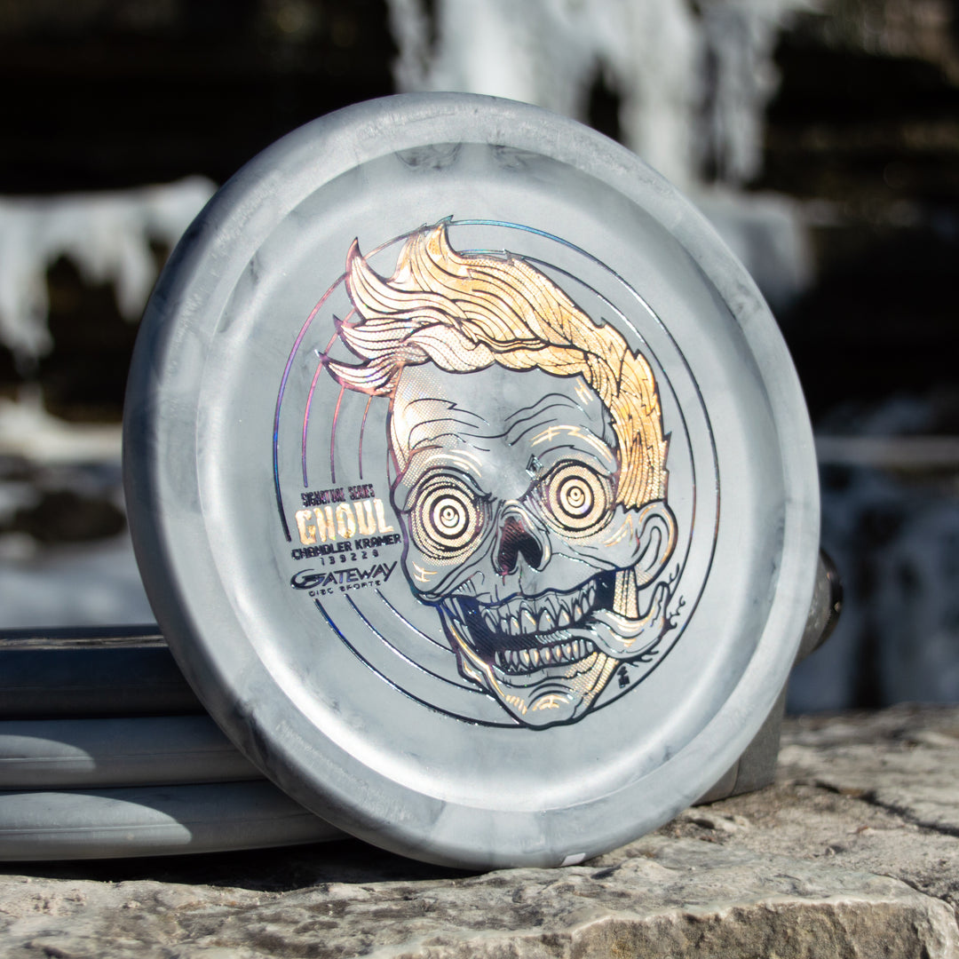 The Chandler's Special Edition Ghoul - Lunar is a gray disc golf disc featuring an illustrated zombie-like face, yellow hair, and exaggerated features—ideal for Power Throwers wanting a stable overstable approach disc, shown on a stone surface outdoors.