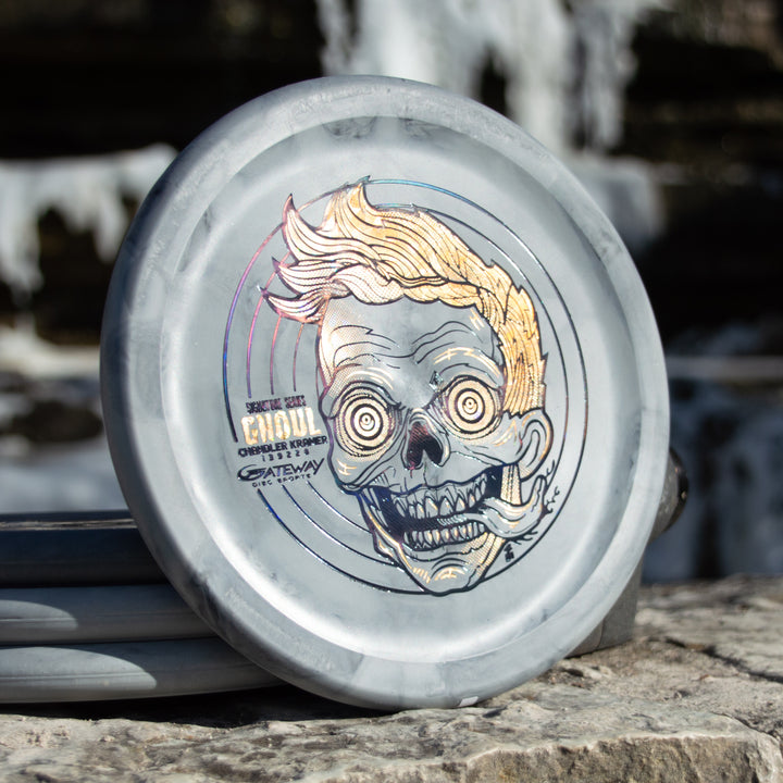 The Chandler's Special Edition Ghoul - Lunar is a gray disc golf disc featuring an illustrated zombie-like face, yellow hair, and exaggerated features—ideal for Power Throwers wanting a stable overstable approach disc, shown on a stone surface outdoors.