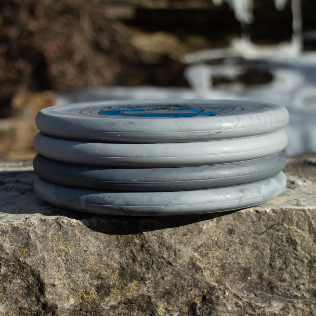 A stack of four Chandler's Special Edition Ghoul - Lunar gray discs, perfect for Power Throwers and offering Forehand Comfort, rests outdoors on a rough stone surface.