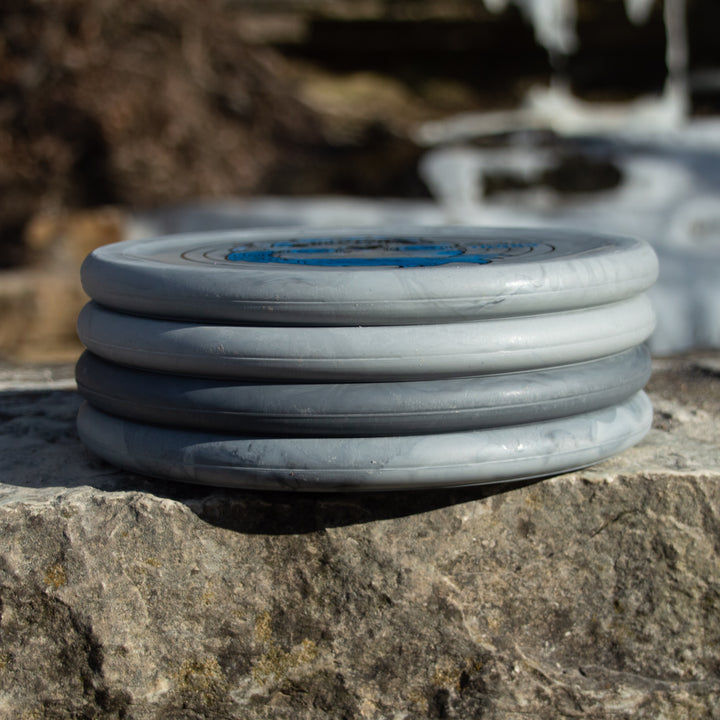 A stack of four Chandler's Special Edition Ghoul - Lunar gray discs, perfect for Power Throwers and offering Forehand Comfort, rests outdoors on a rough stone surface.