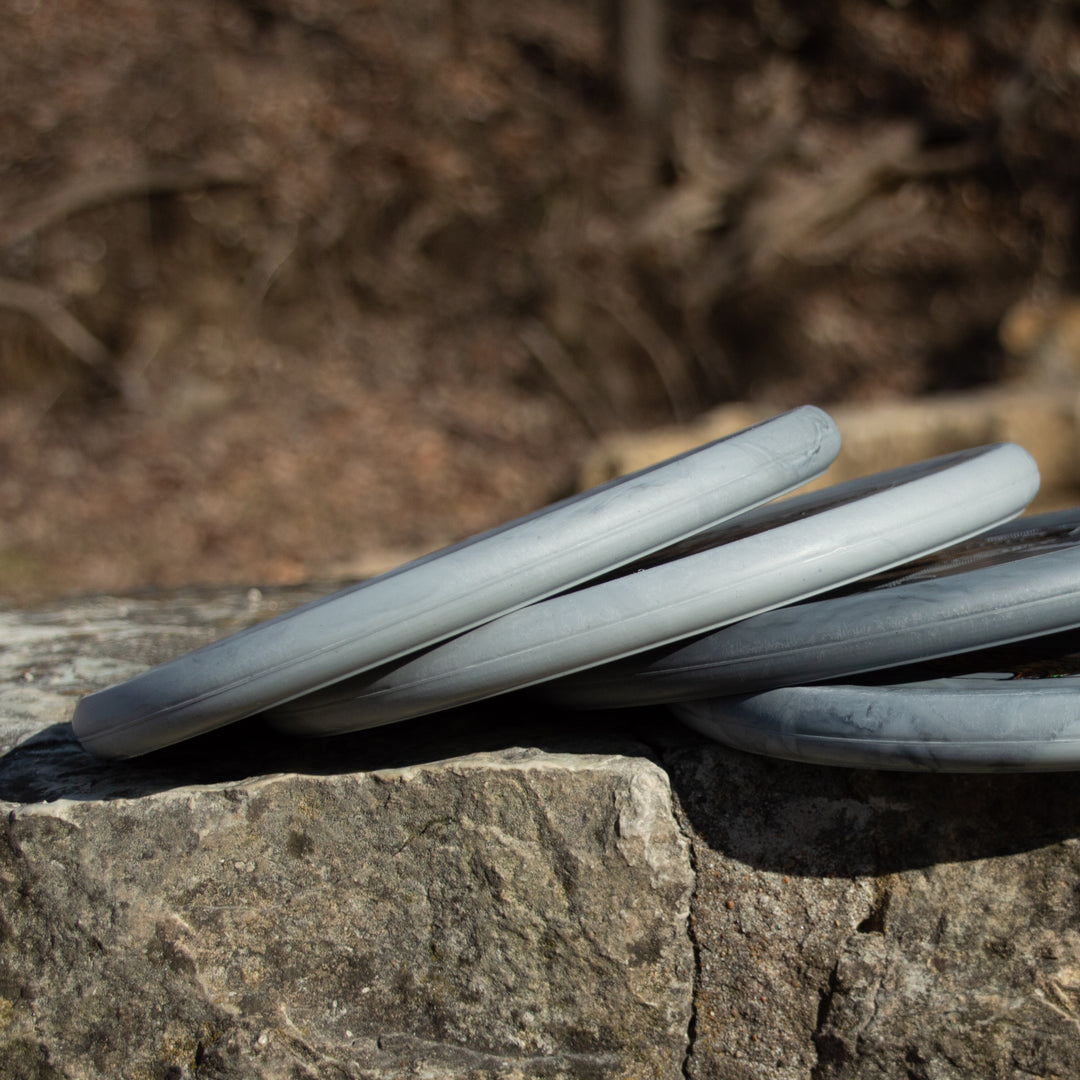 Four gray Chandler's Special Edition Ghoul - Lunar discs are stacked on a large rough stone outdoors, with brown foliage and trees in the background—ideal for power throwers seeking comfortable forehand approach shots.