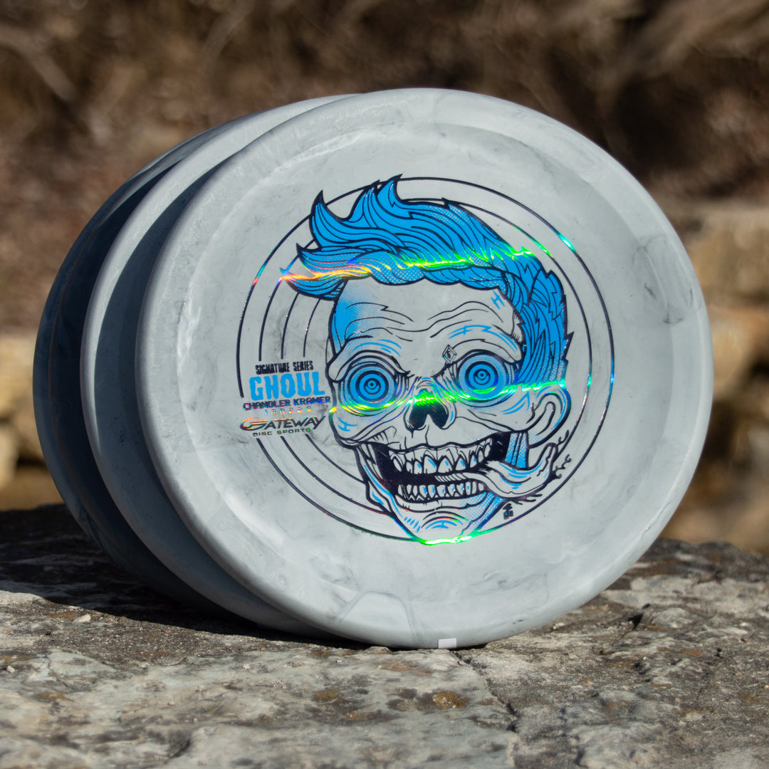 A stack of Chandler's Special Edition Ghoul - Lunar gray disc golf discs, featuring a blue-haired, skull-faced graphic and "GHOUL" text, sits on a stone surface—ideal for Power Throwers wanting a stable overstable approach disc.