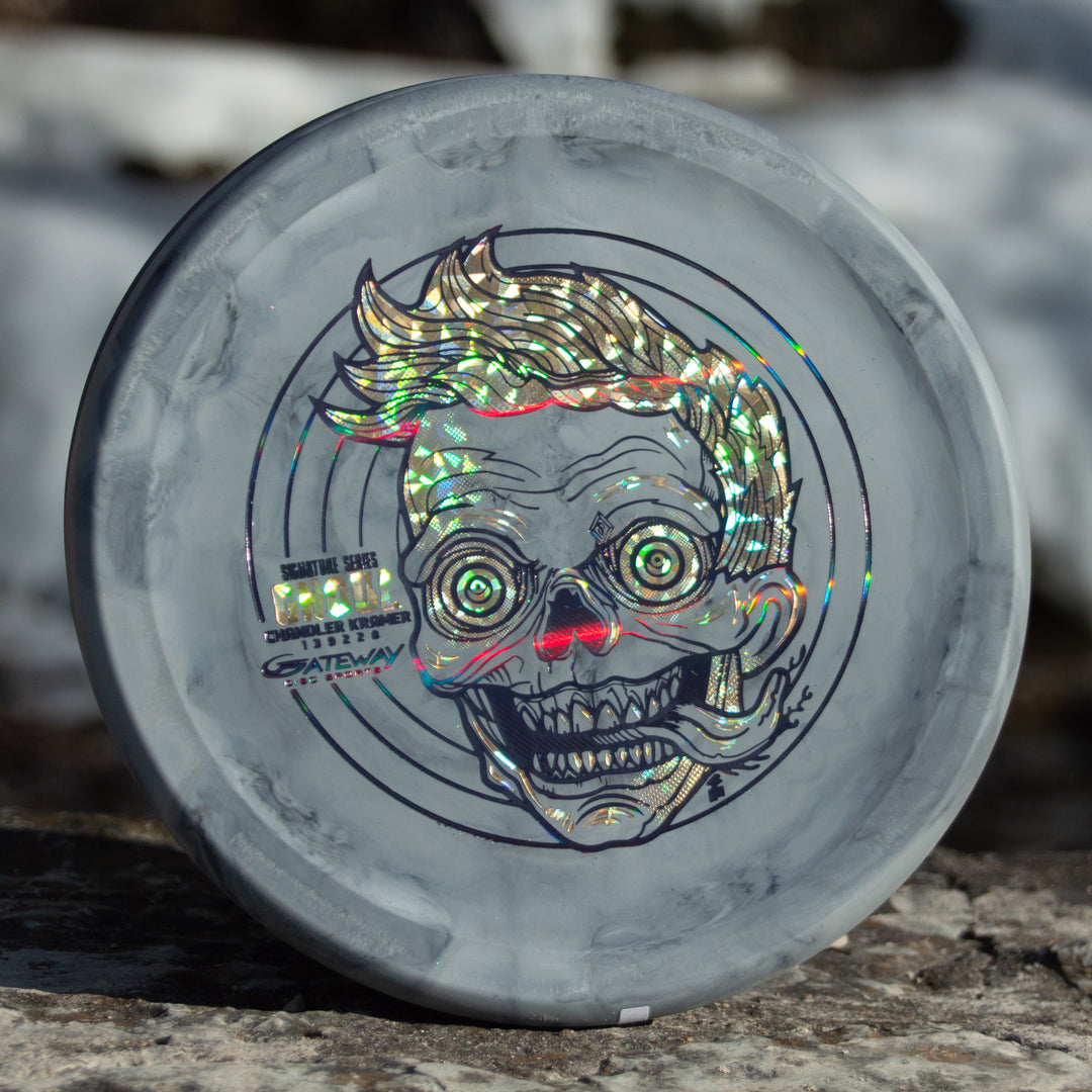 A gray Chandler's Special Edition Ghoul - Lunar, featuring a shiny holographic cartoon skull with exaggerated eyes and hair on the front, rests outdoors on stone—an overstable approach disc ideal for comfortable forehand and power throws.