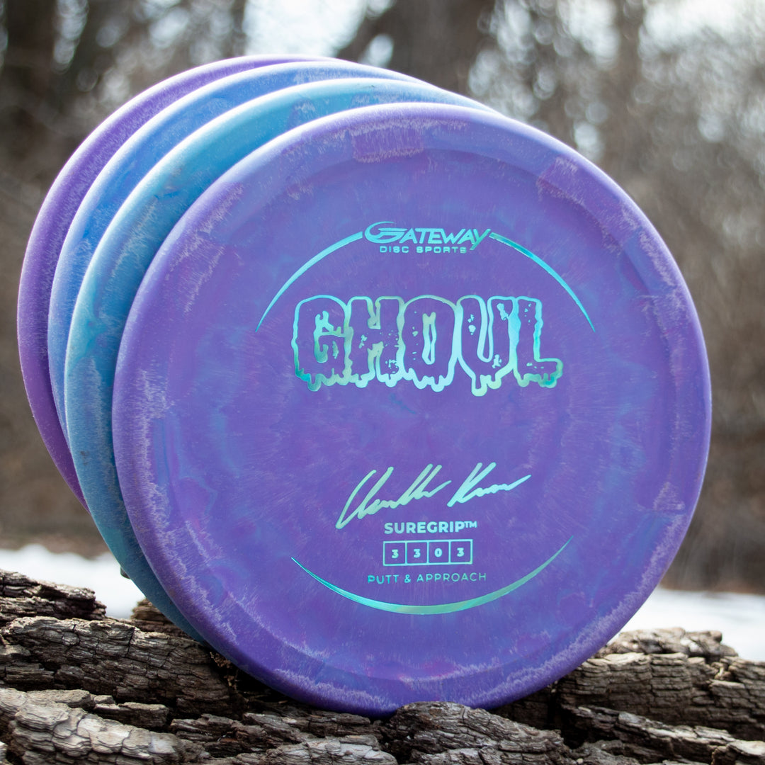 Four Ghoul - Chandler Kramer Signature Series Suregrip 3303 putt and approach discs, in blue and purple, are stacked on a log outdoors against a blurred background of trees.