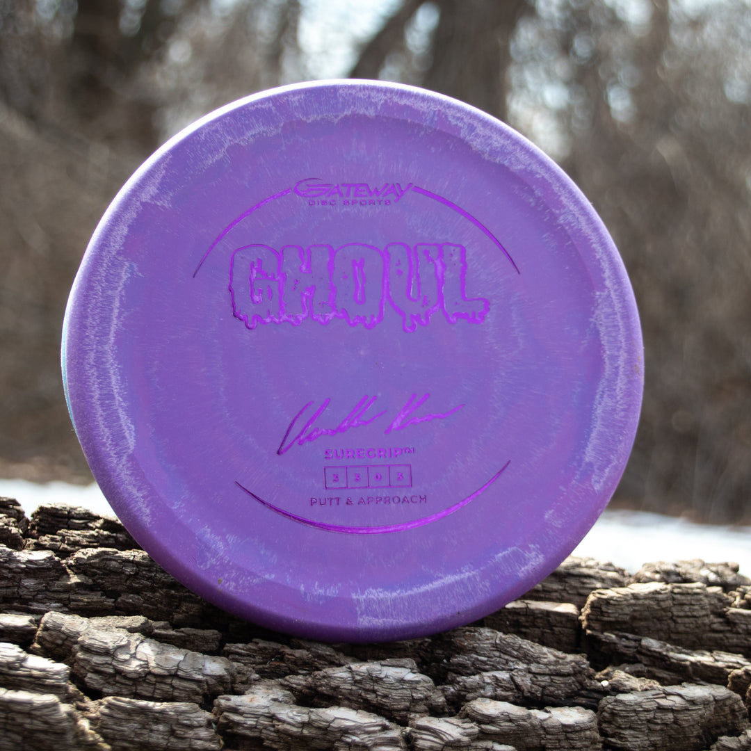 A purple Ghoul - Chandler Kramer Signature Series putter sits on a tree trunk with a blurred natural backdrop, delivering consistent, reliable flight for confident disc golf putts.
