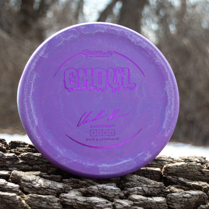 A purple Ghoul - Chandler Kramer Signature Series putter sits on a tree trunk with a blurred natural backdrop, delivering consistent, reliable flight for confident disc golf putts.