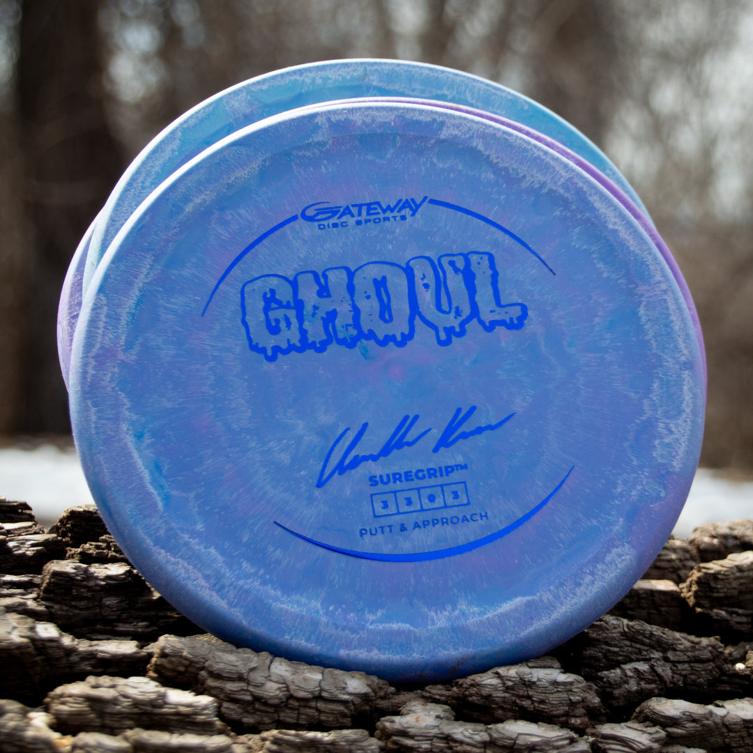 A blue Ghoul - Chandler Kramer Signature Series disc golf putter, designed for stable approaches, lies on rough bark outdoors with a blurred forest in the background.