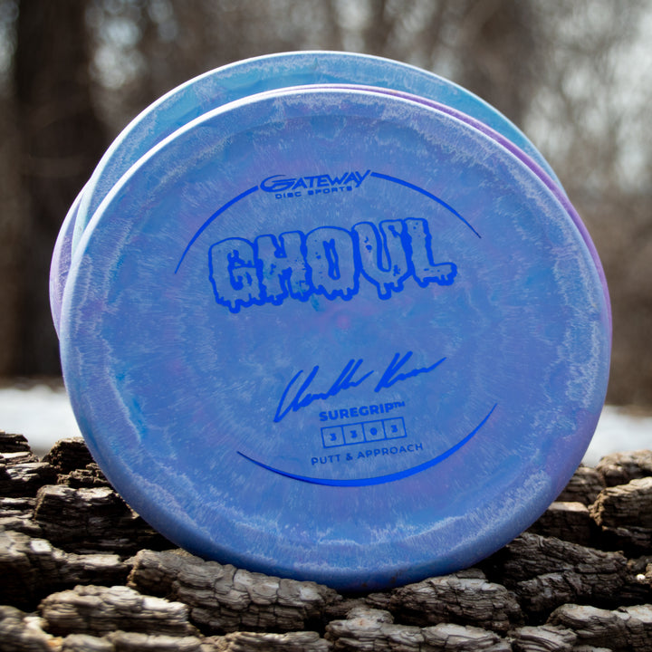 A blue Ghoul - Chandler Kramer Signature Series disc golf putter, designed for stable approaches, lies on rough bark outdoors with a blurred forest in the background.