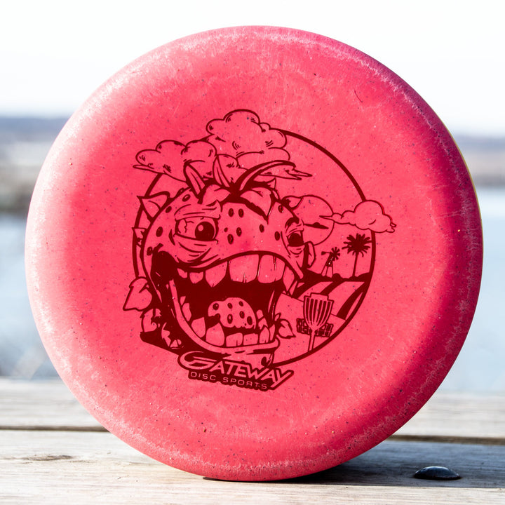 A Wizard Strawberry Hemp disc golf putter, stable for putting and mid-range drives, features a cartoon dinosaur with basket and "Gateway Disc Sports" text, displayed on an outdoor wooden surface.