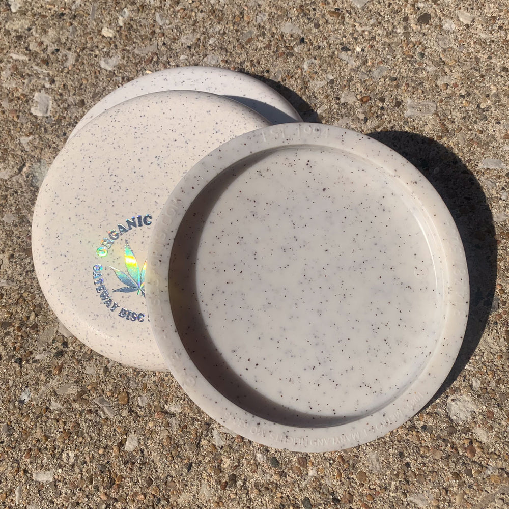 Two speckled white plastic discs, one labeled "Mini (Large) - Hemp," rest on a rough concrete surface in sunlight.