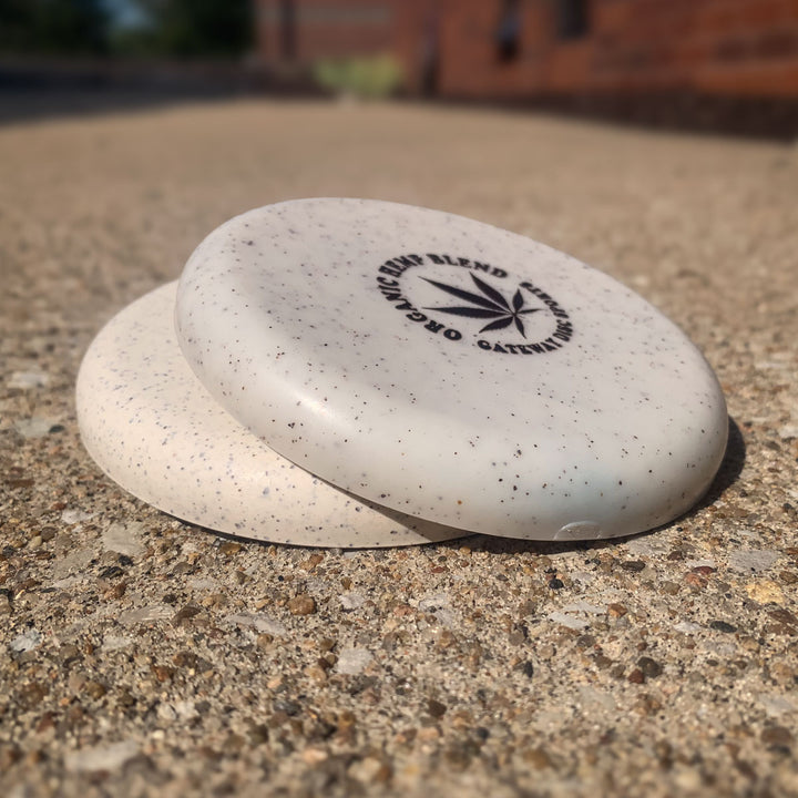 Two Mini (Large) - Hemp speckled white disc golf discs are stacked on a rough concrete surface, with a blurred brick building and outdoor scenery in the background.