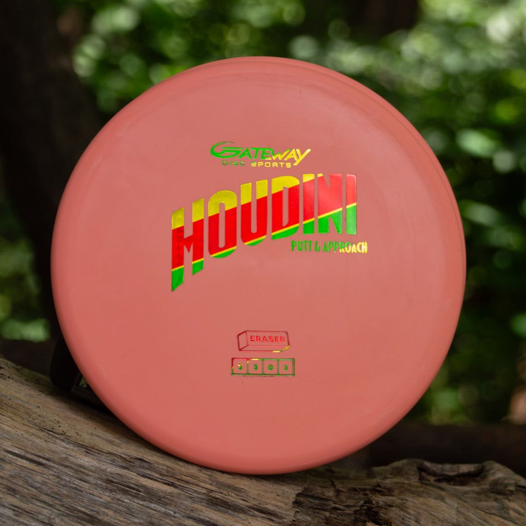 A pink Houdini - Eraser putt and approach disc by Gateway Disc Sports rests on a log, ideal for powerful backhands, with a blurred green forest in the background.