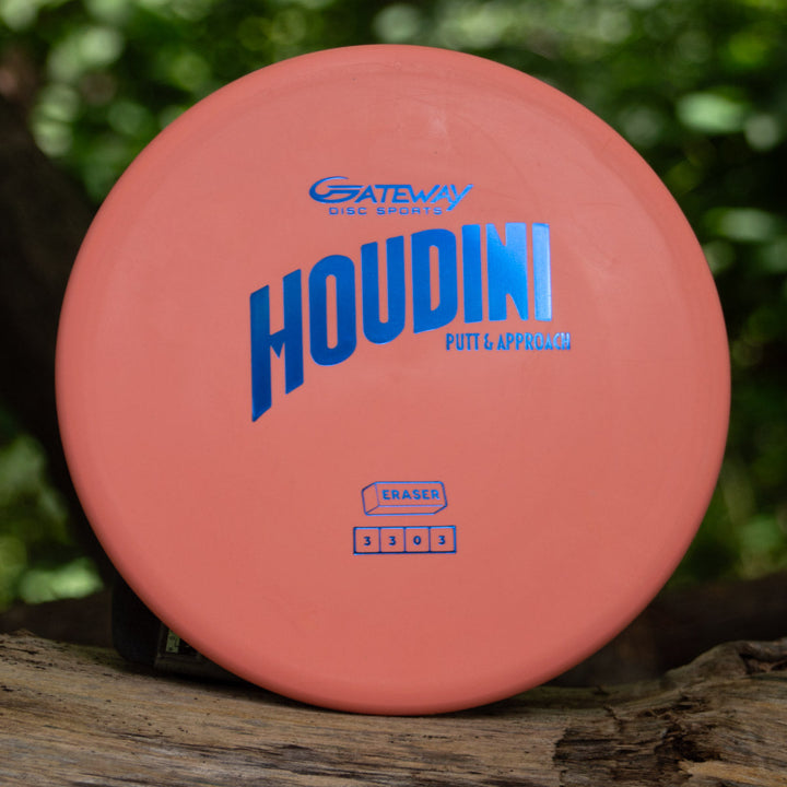 An orange Houdini - Eraser putt and approach disc with blue lettering sits on wood outdoors, ready for smooth putts or power backhands, surrounded by lush green foliage.