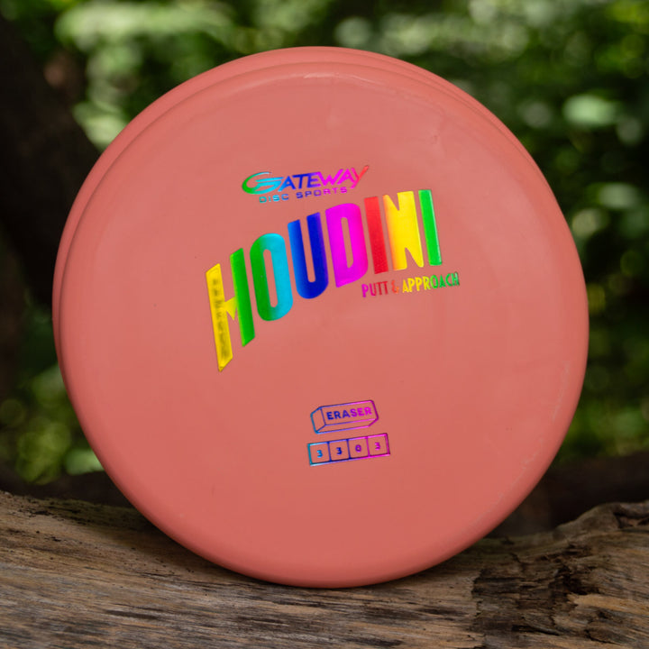 A coral-colored Houdini - Eraser putt and approach disc with holographic lettering sits on a wooden surface outdoors, ready for confident power-grip backhands.