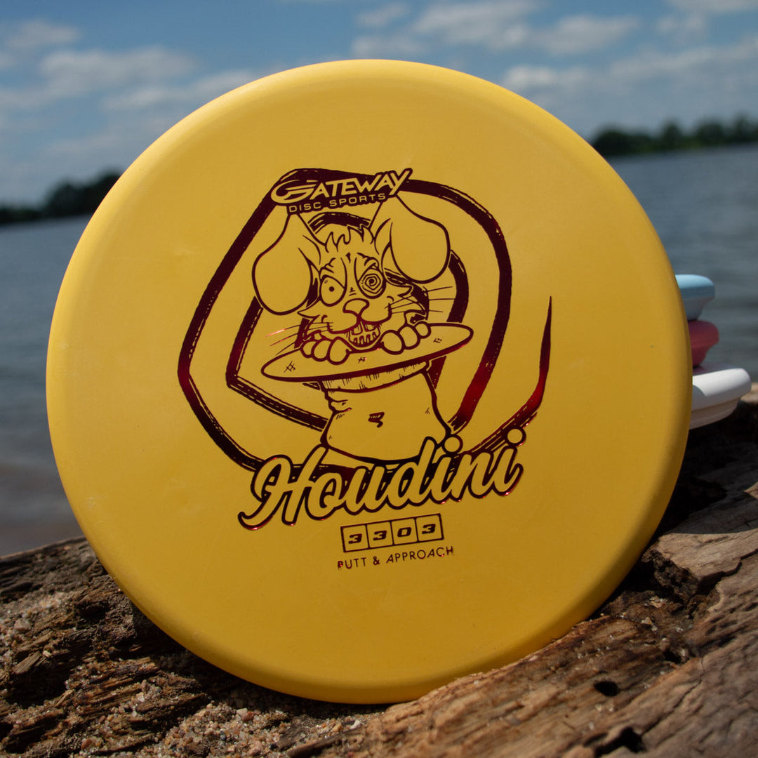 The yellow Houdini - SureGrip™  disc golf putter features a cartoon rabbit holding a disc, displayed on a log by the water—ideal for powerful backhand throws.