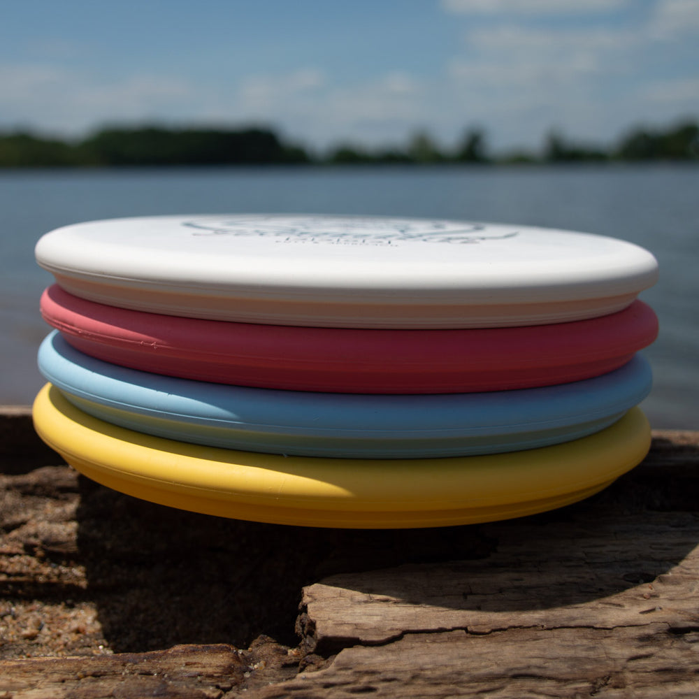 A stack of Houdini - SureGrip™  disc golf discs—ideal for putt and approach shots—rests on a lakeside log beneath a partly cloudy sky, ready for power-gripped backhand throws.