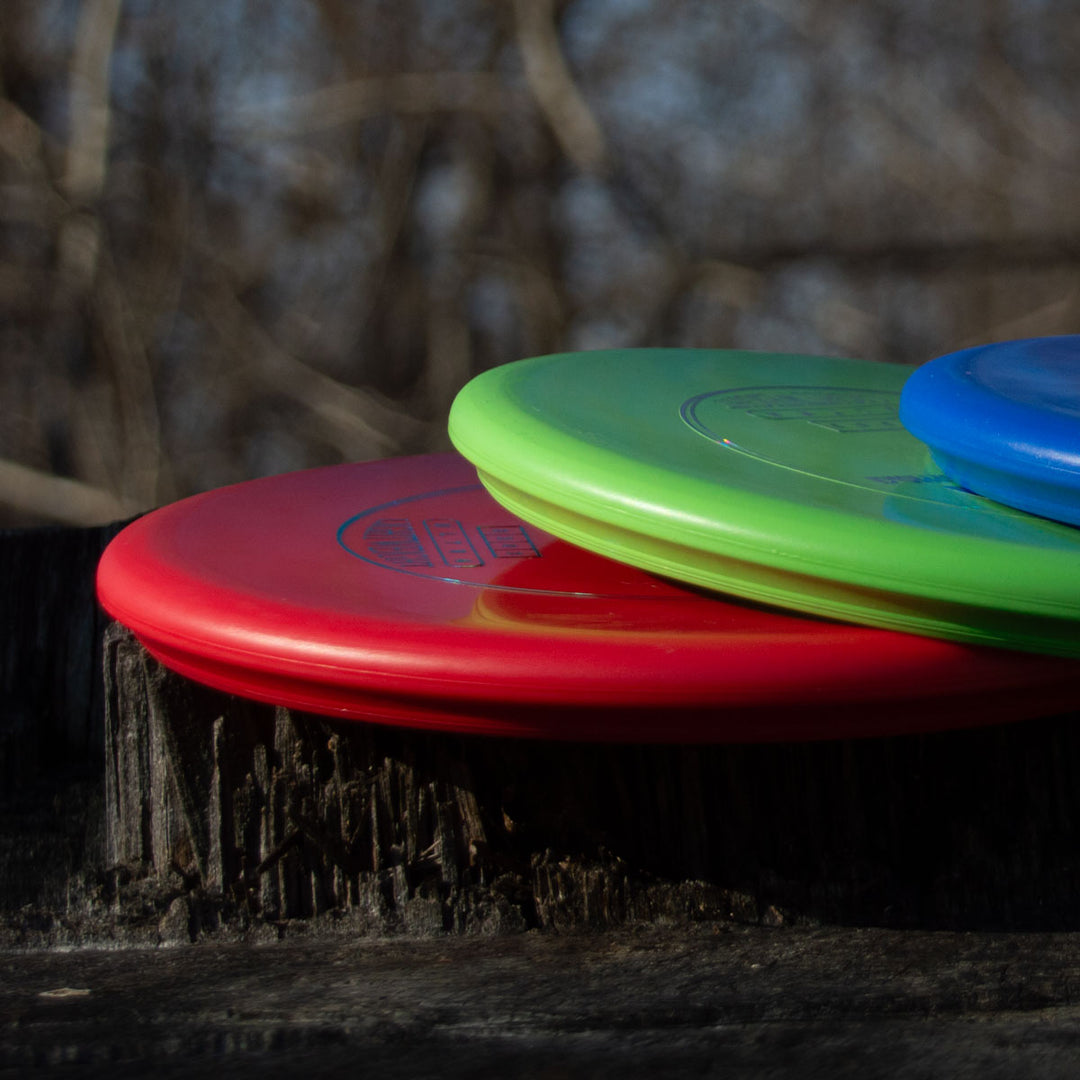 Three Houdini - SureGrip™ Ultra Lightweight putt and approach discs in red, green, and blue are stacked on a tree stump outdoors, with bare branches blurred in the background.