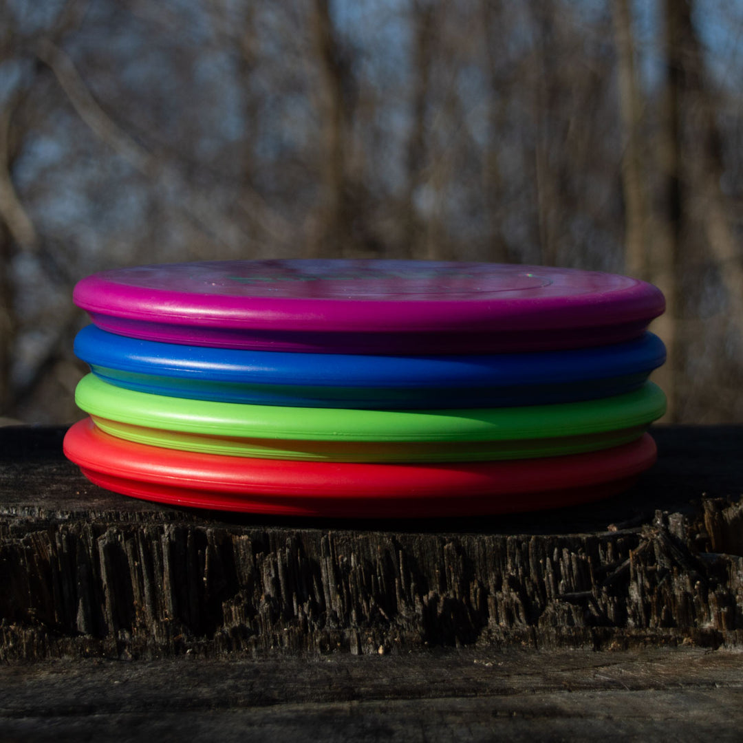 A stack of Houdini - SureGrip™ Ultra Lightweight putt and approach discs in pink, blue, green, and red, each featuring a deep rim, rests on a weathered wooden surface with trees softly blurred in the background.