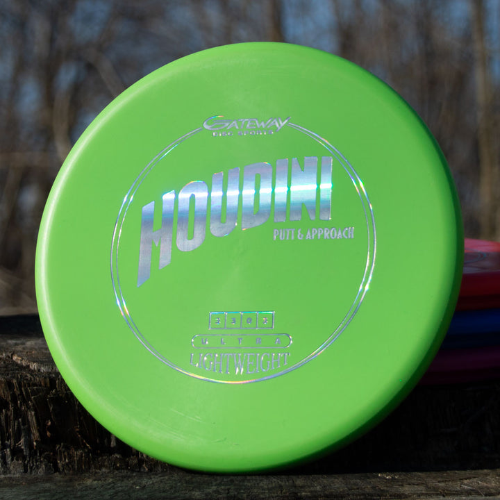 A green Houdini - SureGrip™ Ultra Lightweight putt and approach disc is shown on a wooden surface outdoors with trees and discs blurred behind it. Its deep rim design provides confidence for your throws.