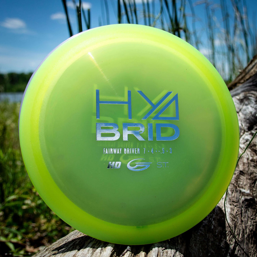 A yellow-green Hybrid - HD fairway driver in Hyper Diamond blend sits on a log outdoors, with tall grass and a lake in the background.