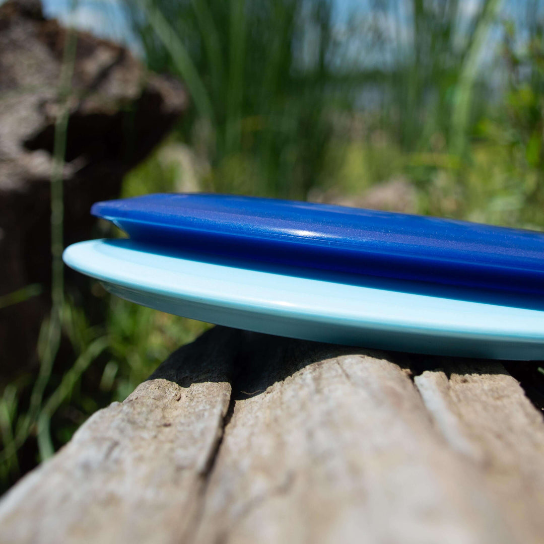 Two plastic flying discs, one blue and one light blue, are stacked on a weathered log outdoors. These Hybrid - HD discs feature the Hyper Diamond blend, offering players a premium fairway driver with excellent grip and durability.