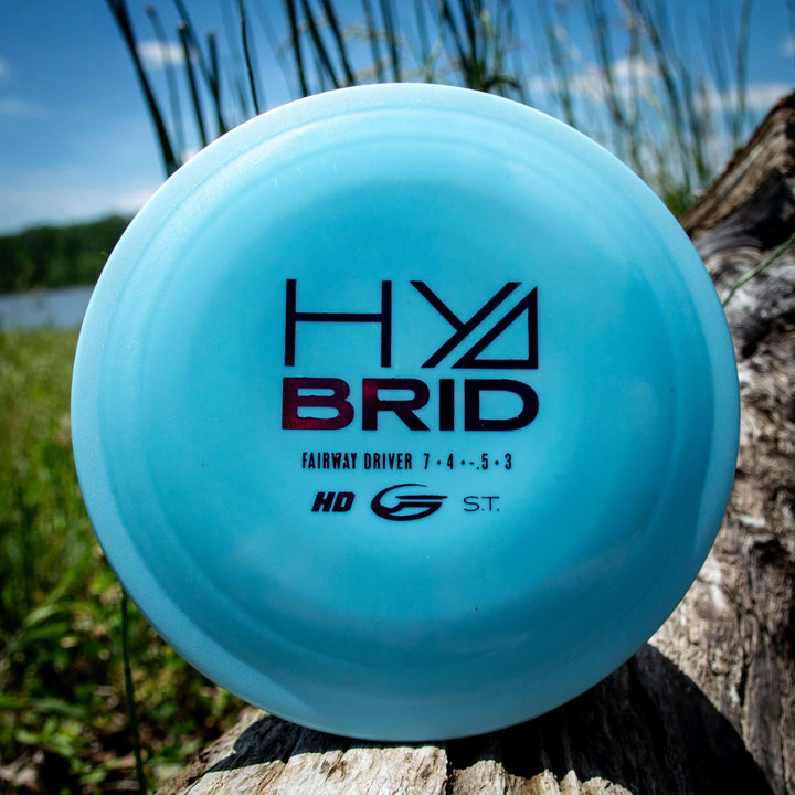 A blue Hybrid - HD disc fairway driver in Hyper Diamond blend, printed with "HYBRID" and flight numbers 7, 4, -5, 3, rests on a log outdoors with grass and water in the background.