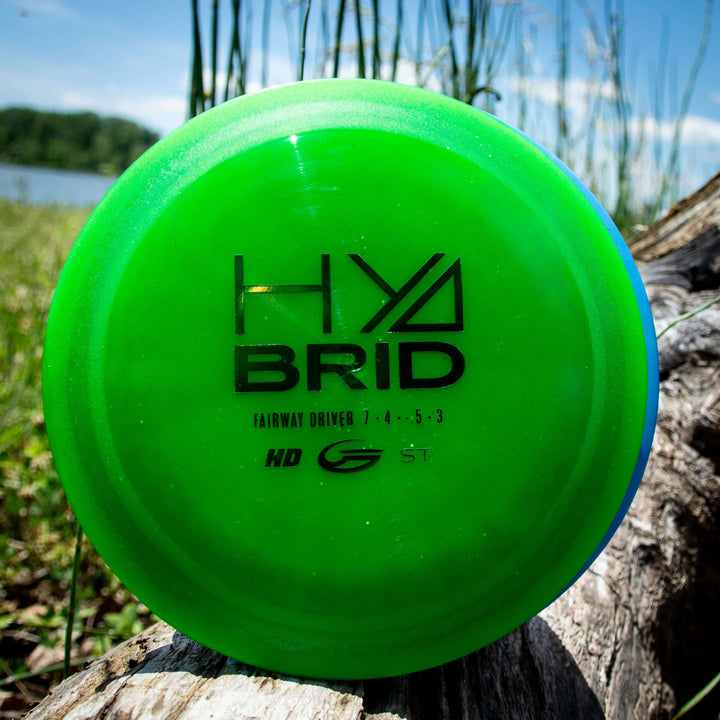 A green Hybrid - HD disc fairway driver in Hyper Diamond blend rests against a log outdoors, with tall grass and water behind it—perfect for your next round.