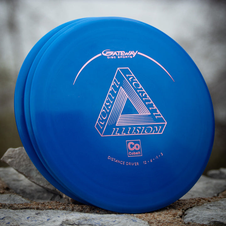 A blue Illusion - Cobalt distance driver by Gateway Disc Sports, made with easy-to-throw cobalt blend plastic, sits on a stone surface outdoors.