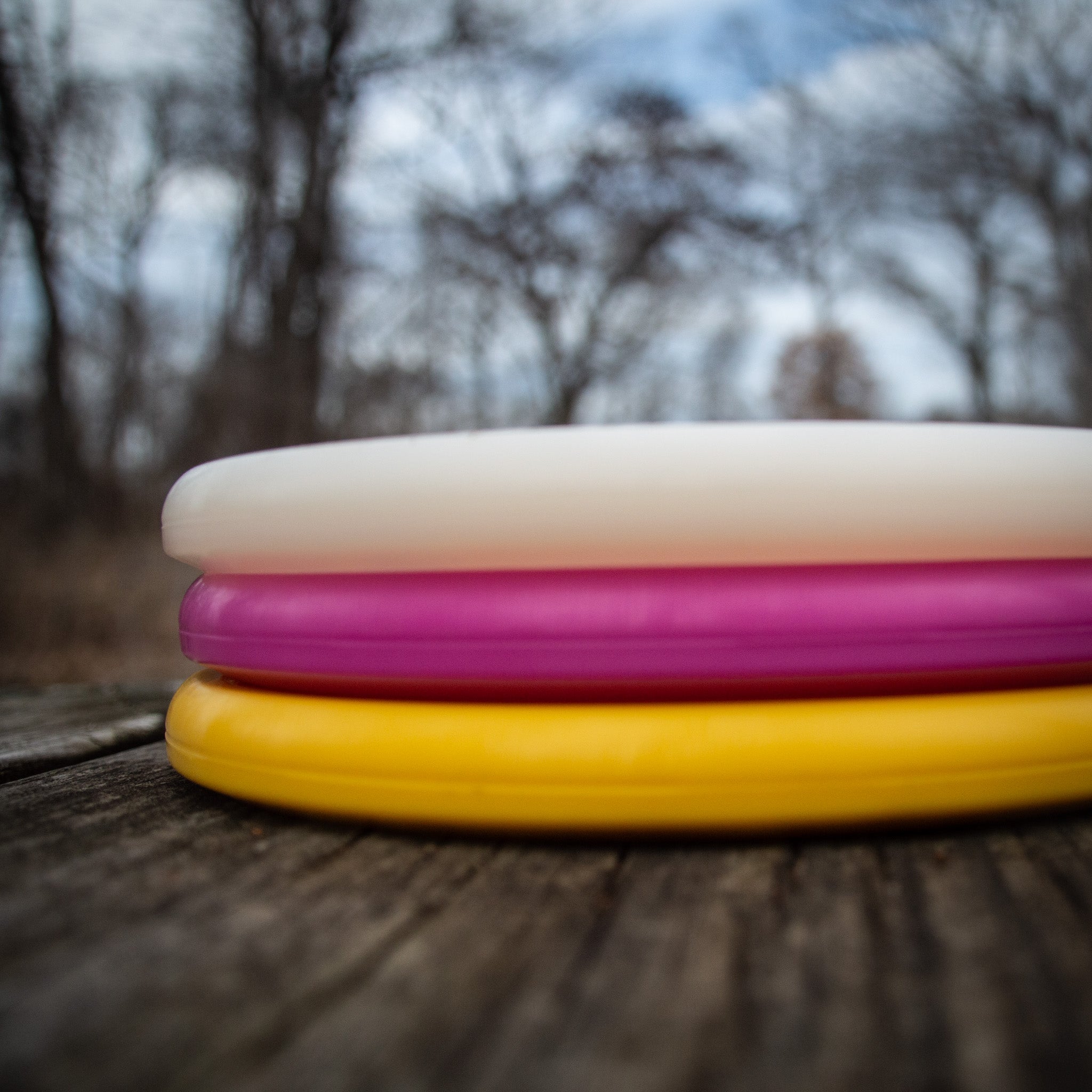 Three Warspear - NXT - Ivan Marston Artist Series discs—white, pink, and yellow—made from a durable plastic blend are stacked on a wooden surface outdoors with bare trees and a cloudy sky in the background.