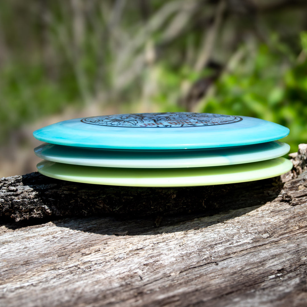 Three vibrant Jon Borzick's Ether - Color Superglow distance drivers by Gateway Disc Sports, made with durable NXT material, stacked on a log outdoors against a blurred background of greenery and branches.
