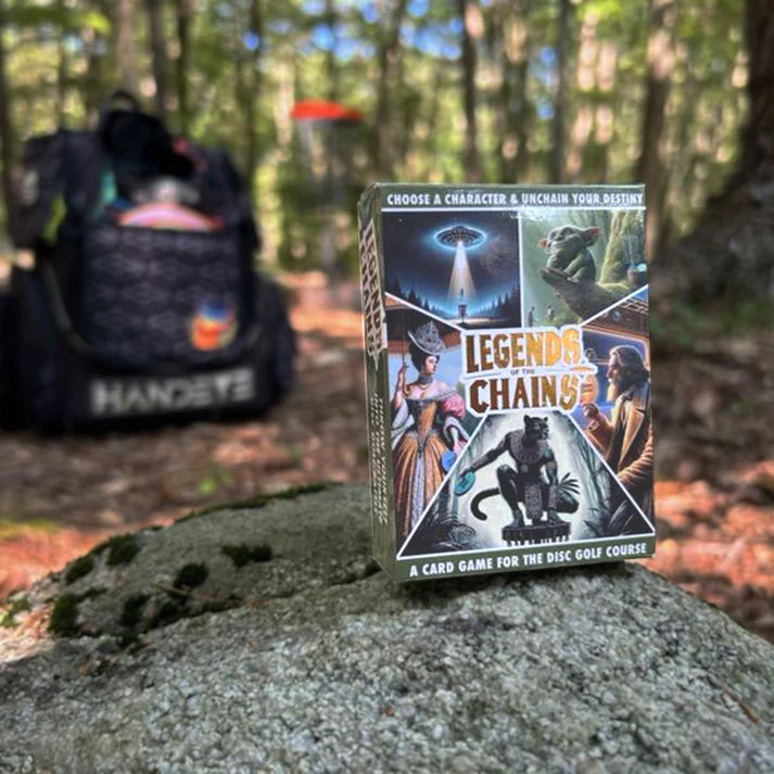 A box of "Legends of the Chains: On-Course Disc Golf Game" rests on a rock in the woods, with character cards visible and a disc golf bag and flying disc blurred in the background.